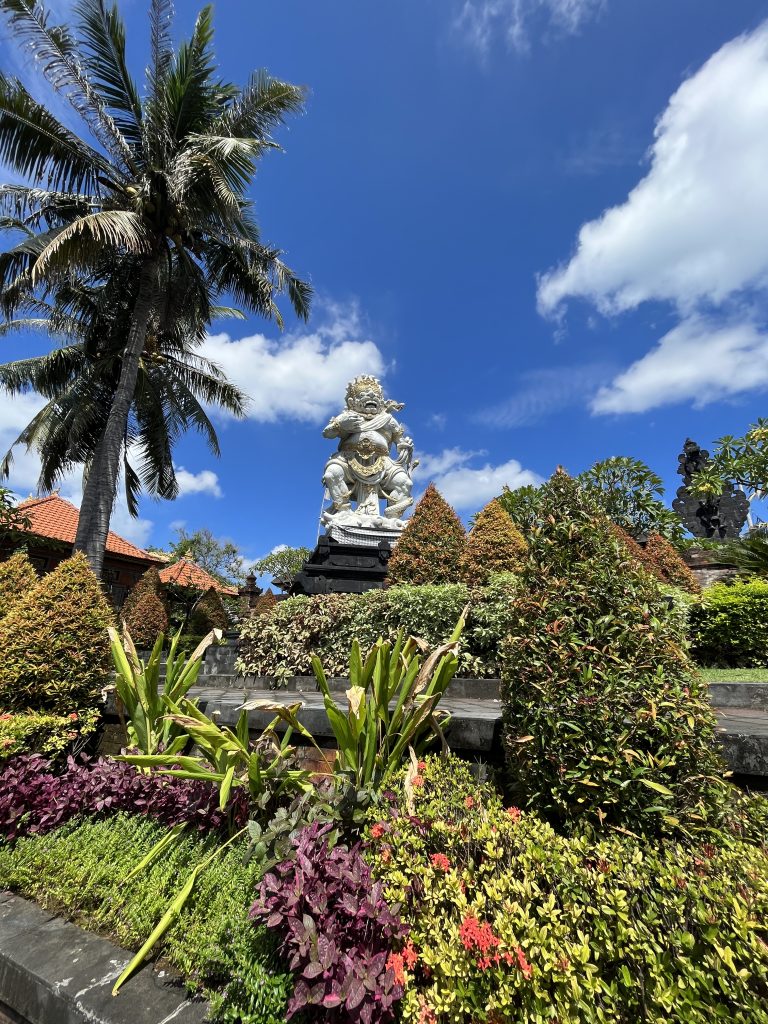 A statue in Bali, surrounded by a garden, photography against a partially cloudy blue sky. Large palm trees can be seen next to the statue.