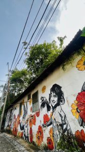 View larger photo: A painted mural on an outside wall depicting a person, flowers, a dog and a bird. The sky overhead is blue with fluffy clouds and electric wires run through it.