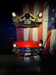 A wall with an American flag draped over it. The front bumpers of a yellow 1950 Studebaker and a red 1957 Corvette are mounted on the wall.
