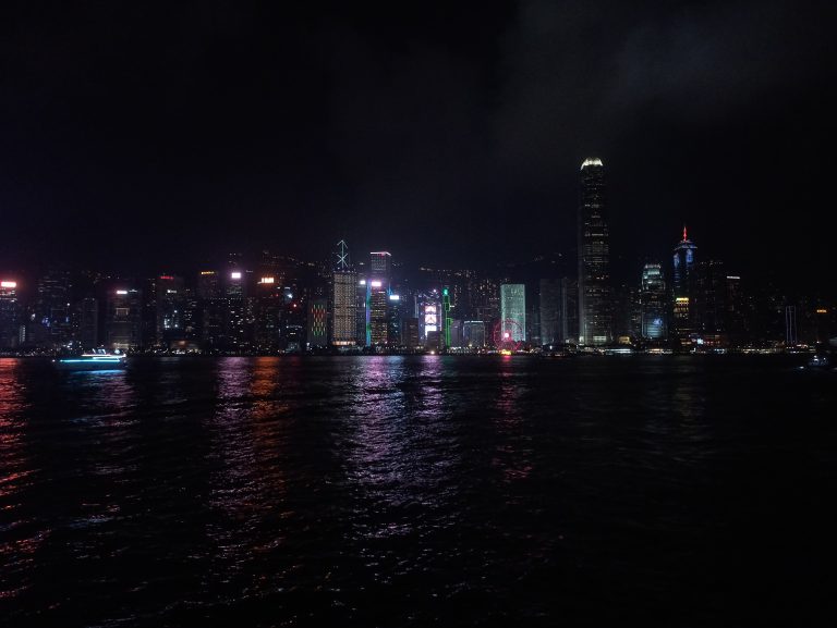 Hong Kong nighttime cityscape, illuminated by colorful lights, reflecting on the water, with a dark sky overhead.