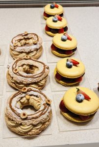 A display of assorted pastries: on the left, there are circular pastries with a cream filling and hazelnut toppings, some dusted with powdered sugar. On the right, there are yellow macaron-like pastries filled with cream and fresh fruit, including strawberries and blueberries.