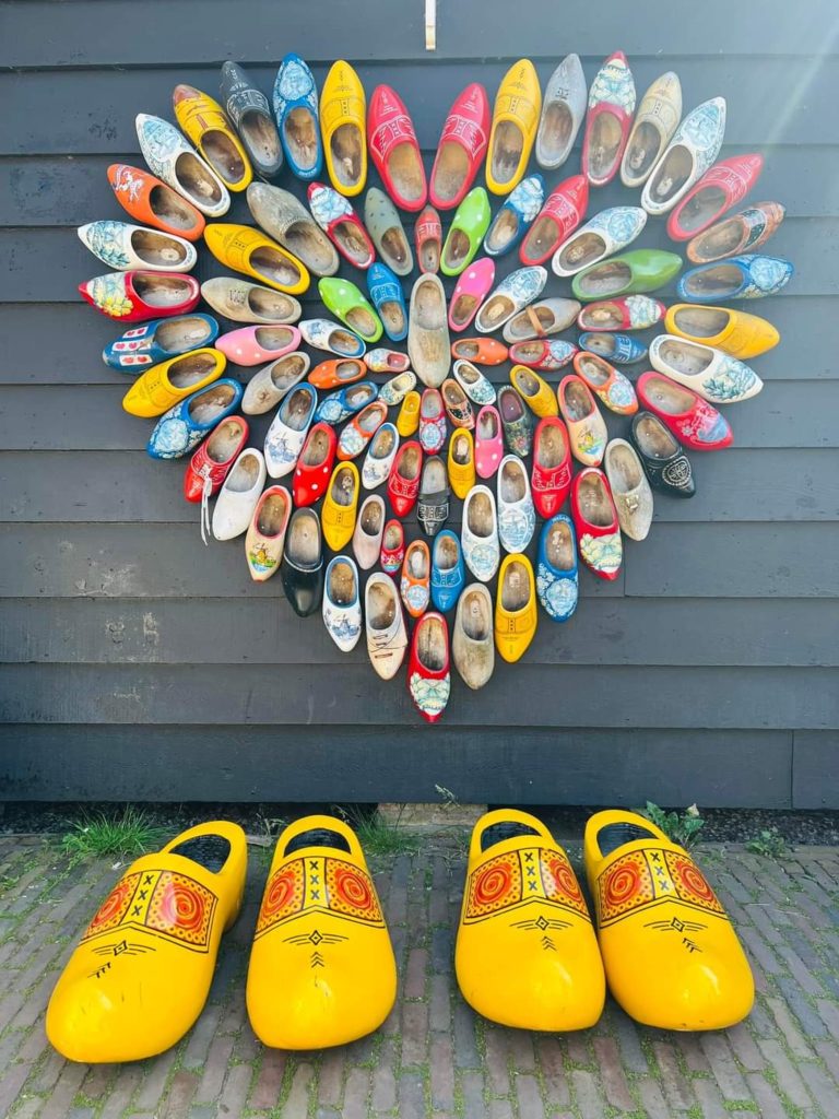 A large number of wooden shoes arranged in the shape of heart on the exterior wall of a wooden building at Zaanse Schans in The Netherlands. Four giant clogs can be seen on the ground below the heart.