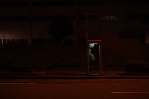 View larger photo: A phone booth on the sidewalk next to a road, at night. Red light illuminates the street. A fence and some buildings can be seen in the background.