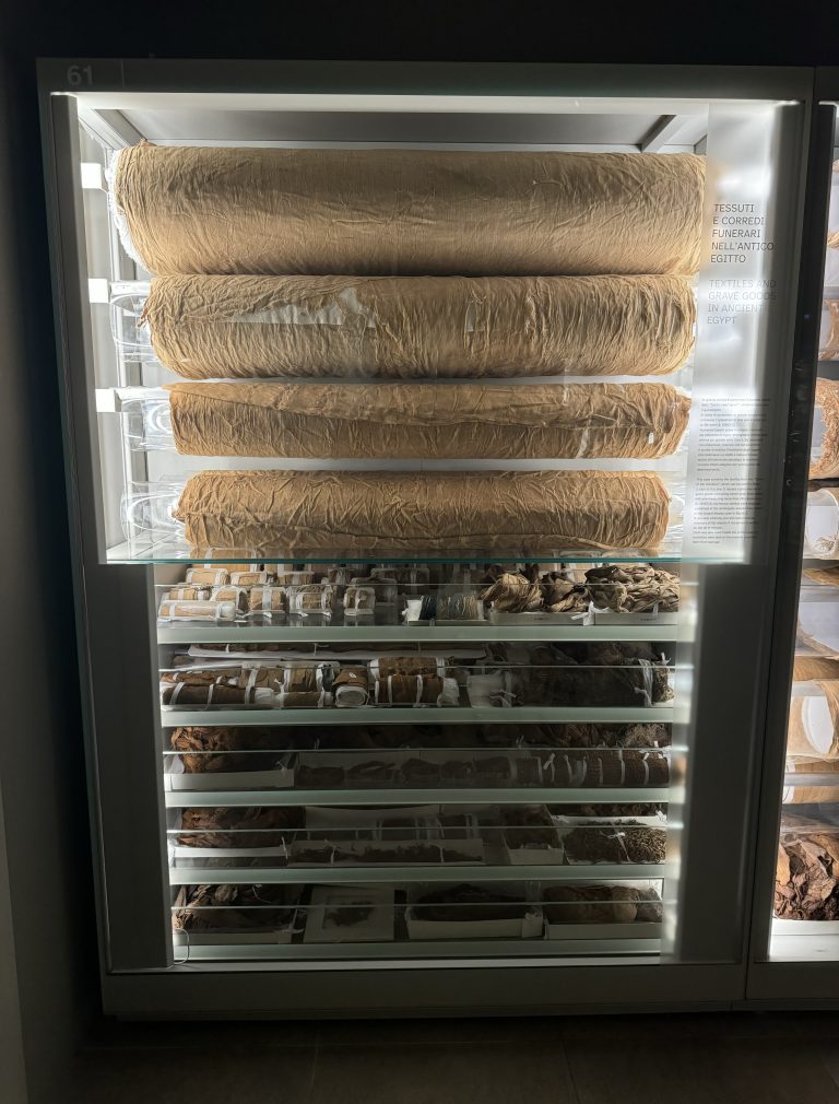 A sealed display case with textiles that were discovered as grave goods in the “Tomb of the Unknown”. Seven large rolls of linens are on the top with other assorted pieces below. This collection is found at the Museo Egizio in Turin, Italy.