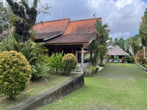 A traditional Balinese wooden house with a red-tiled roof, surrounded by lush greenery and a garden path leading to a gazebo.
