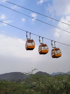 Three orange cable cars suspended mid-air against a backdrop of mountains and greenery, with a cloudy blue sky above.