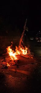 A fire burns in the middle of a street at night. Flames engulf a pile of debris including a corrugated sheet and pieces of wood. The surroundings are dimly lit, with a wall and a parked vehicle in the background.



