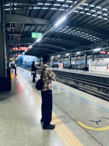 A young man waiting for a train on the platform of a metro station. Several other passengers can be seen in the background, out of focus.