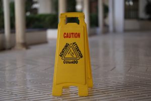 A yellow caution sign placed on a tiled floor indicating a wet or slippery surface. The sign bears the words "CAUTION," "ACHTUNG," "ATTENTION," and "CUIDADO" above a symbol of a person slipping.