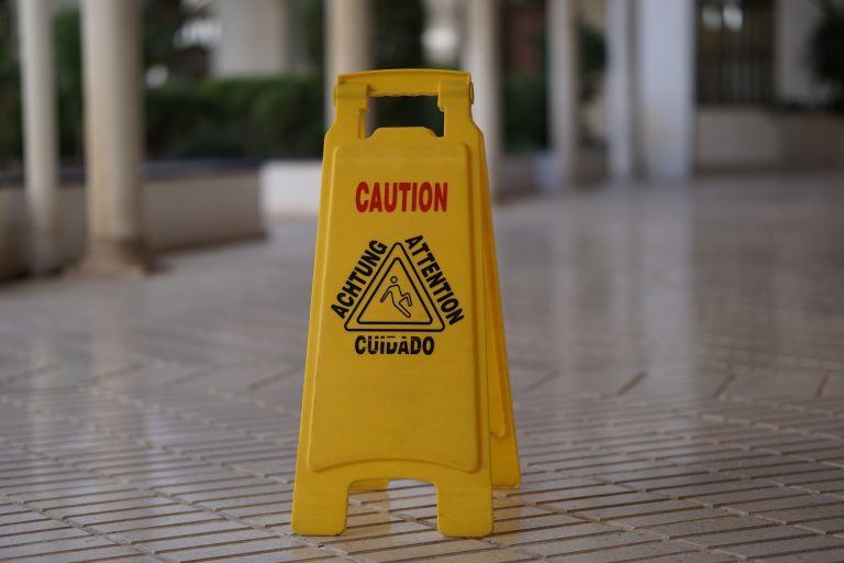 A yellow caution sign placed on a tiled floor indicating a wet or slippery surface. The sign bears the words “CAUTION,” “ACHTUNG,” “ATTENTION,” and “CUIDADO” above a symbol of a person slipping.