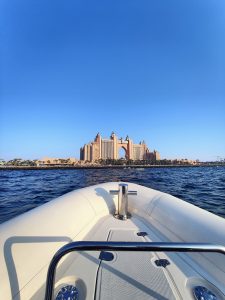 The view from a boat's bow looking towards the iconic Atlantis hotel, with its large central archway, set against a clear blue sky and calm ocean waters.