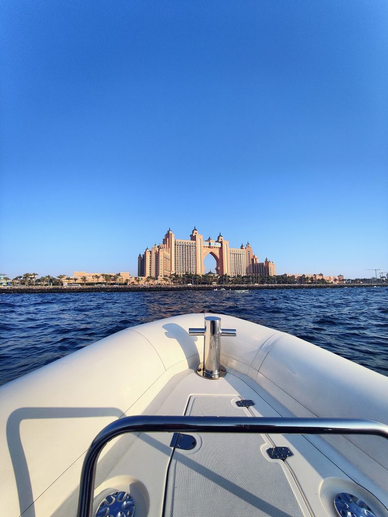 The view from a boat’s bow looking towards the iconic Atlantis hotel, with its large central archway, set against a clear blue sky and calm ocean waters.