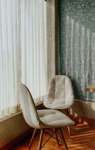 Two white cushioned chairs positioned near a large window with sheer white curtains allowing natural light to enter the room.