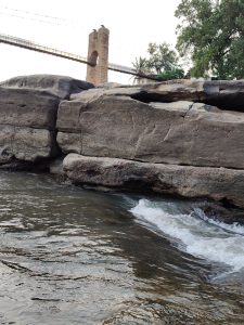 Flowing water of Gokak falls, above large rocks there's a hanging bridge with some brick pillars. 