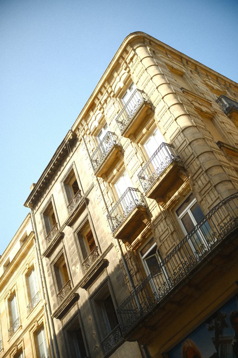 A view of a classic European-style building with ornate wrought iron balconies and tall windows, bathed in sunlight against a clear blue sky.