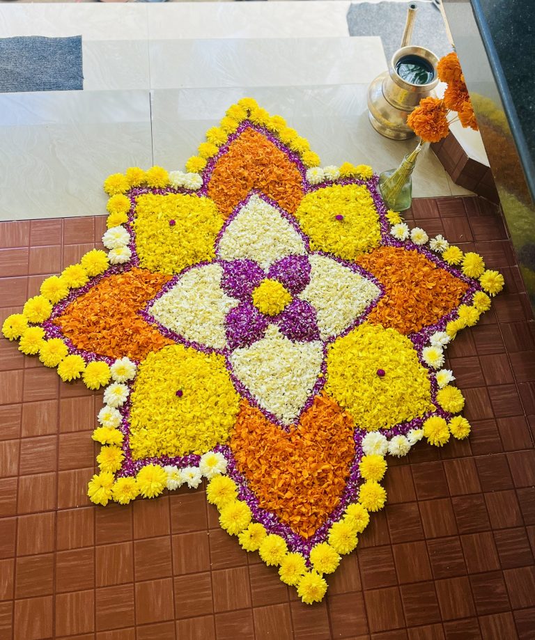 An intricate flower carpet (Pookkalam) on a tile floor near some steps.