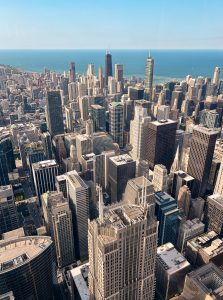 View larger photo: Aerial view of downtown chicago with a dense cluster of skyscrapers and high-rise buildings, extending towards the coastline of lake michigan under a clear blue sky.