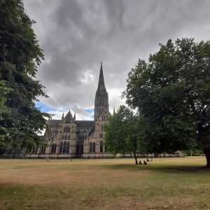 Salisbury Cathedral taken from the Theological College in The Close, in Wiltshire, England. Dark rain clouds surround the top of the spire, with blue sky coming through behind. The Cathedral is set in a long lawn with trees.