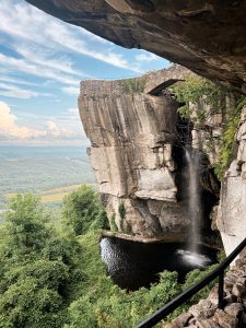 A scenic view of a rocky cliff with a waterfall cascading into a small pool below, surrounded by lush green vegetation. There is a stone bridge at the top of the cliff, and a sign with the text "Lover's Leap" visible on the cliff edge. The background includes a panoramic view of rolling hills under a partly cloudy sky.