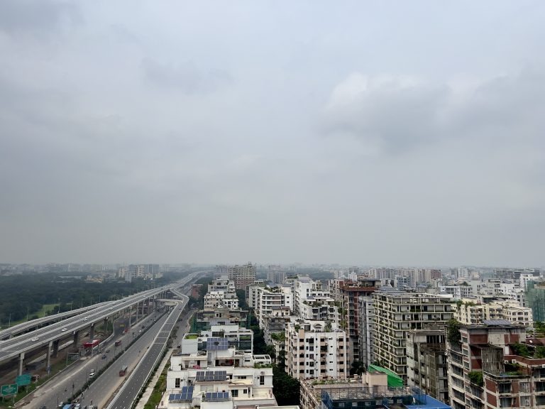 View of Dhaka, Bangladesh, taken from an elevated position. The express highway is visible on the left. The sky is cloudy and gray.