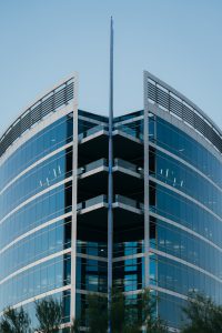 View larger photo: Modern office building with a V-shaped glass facade, multiple balconies, and a central vertical pole, featuring sleek architectural lines and reflective windows against a clear sky backdrop, surrounded by greenery.