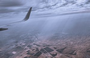 View larger photo: View from the airplane window with a plane wing on the left, and a cloudy blue sky. The ground shows farms like a patchwork quilt below.