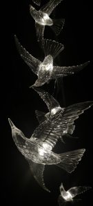 View larger photo: A vertical arrangement of illuminated crystal glass bird sculptures against a dark background, creating the appearance of birds in flight.