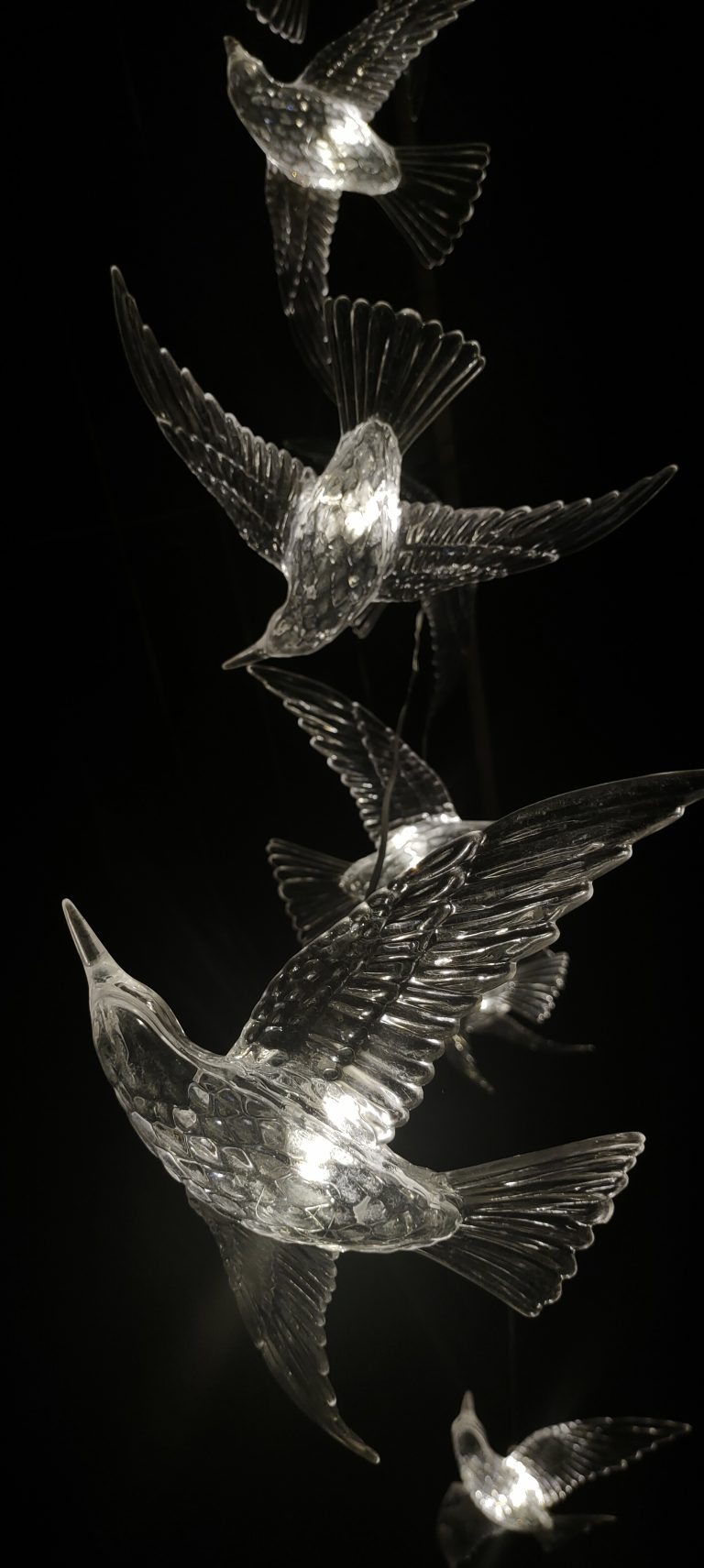 A vertical arrangement of illuminated crystal glass bird sculptures against a dark background, creating the appearance of birds in flight.