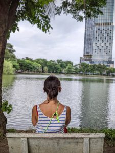 View larger photo: A person sitting on a bench by a lake in Lumphini Park, Bangkok, overlooking a mix of greenery and city skyscrapers in the distance.