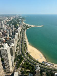 View larger photo: Aerial view of a the chicago cityscape alongside lake michigan and its sandy beaches. Tall buildings and a winding road line the coast, with lush green trees interspersed throughout the city. The sky is clear and blue, and small boats can be seen in the distance on the water.