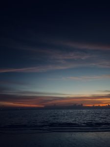 Orange stratus clouds low on an evening horizon. Stars above, water in the foreground.