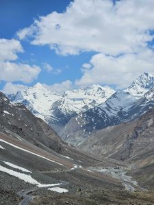 View larger photo: A breathtaking view of snow-capped Himalaya mountains under a blue sky with fluffy clouds, with a winding road snaking through the rocky valley below.