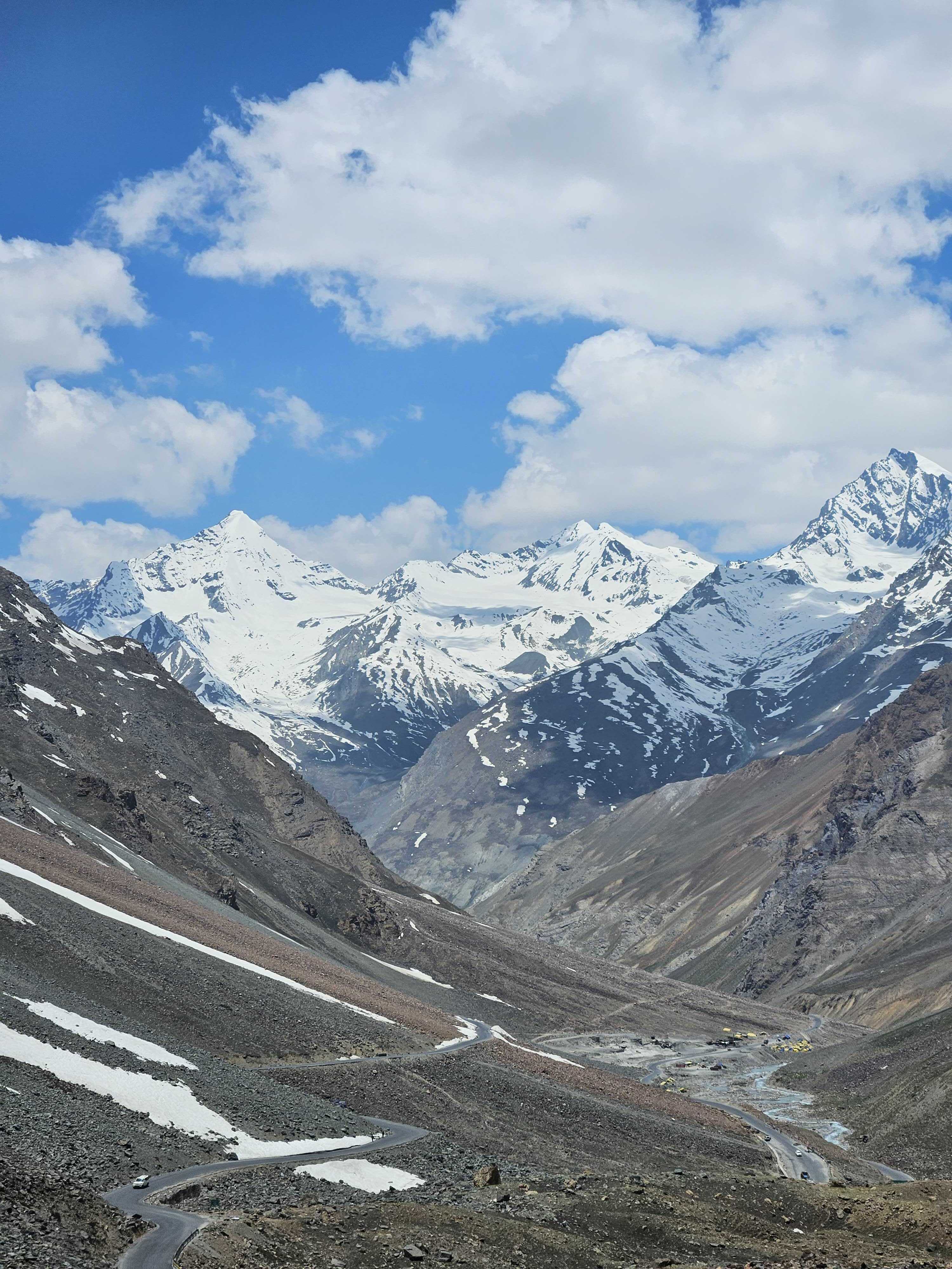 A breathtaking view of snow-capped Himalaya mountains under a blue sky with fluffy clouds, with a winding road snaking through the rocky valley below.