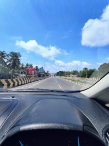 A bright, clear day on a wide road with two lanes in each direction, separated by a yellow and black divider. The view from inside a vehicle shows the road stretching ahead under a blue sky with scattered clouds. Palm trees and billboards line the peaceful roadside.