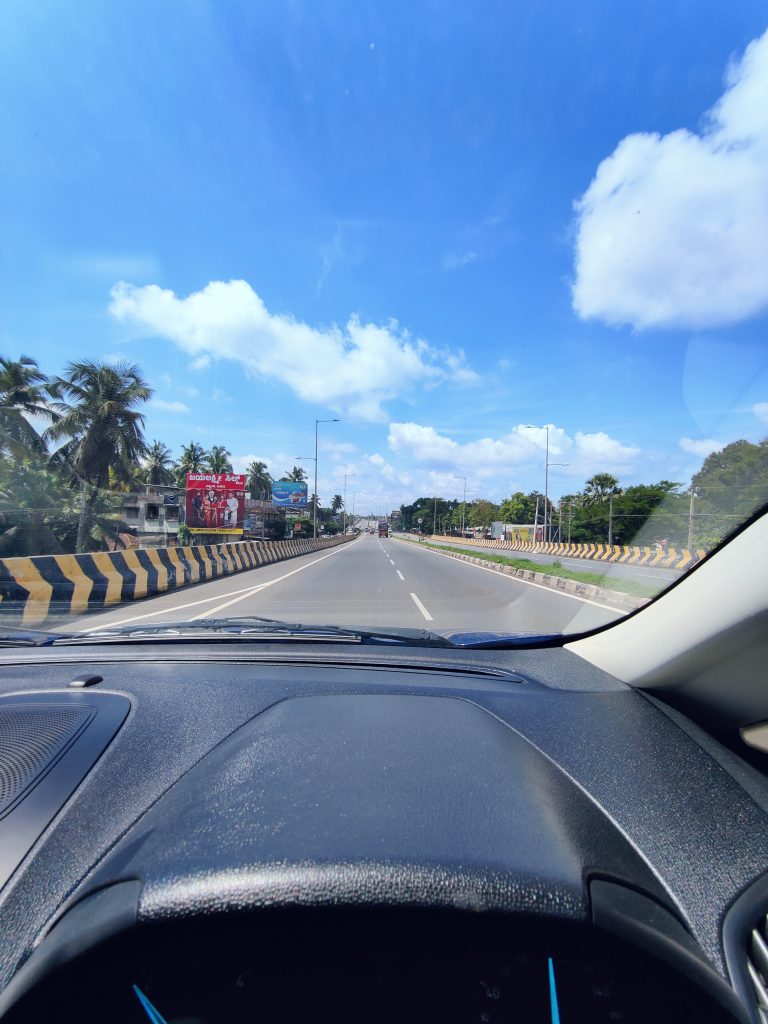 A bright, clear day on a wide road with two lanes in each direction, separated by a yellow and black divider. The view from inside a vehicle shows the road stretching ahead under a blue sky with scattered clouds. Palm trees and billboards line the peaceful roadside.