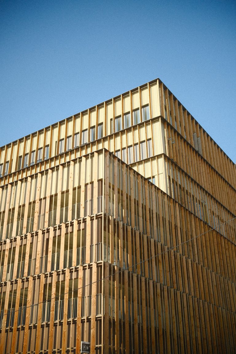 A modern building with vertical wooden slats covering its exterior, featuring large windows, set against a clear blue sky.