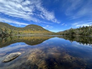 A scenic view of a tranquil lake (Lonesome Lake in New Hampshire) surrounded by mountains covered in autumn foliage. The glass-like clear water of the lake reflects the blue sky and clouds above, along with the neighboring forested hills. A large rock is visible in the foreground under the water's surface. The sky is mostly clear with some scattered white clouds.