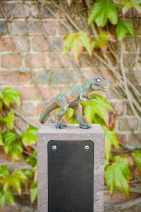 A small plastic dinosaur toy placed on top of a stone post, with a brick wall and green ivy leaves in the background.