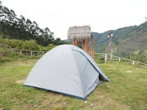 View larger photo: A gray camping tent set up on a grassy area with a wooden structure that has a thatched roof in the background. 