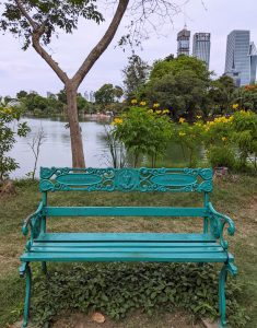 View larger photo: A decorative turquoise colour bench, situated in front of a lake, surrounded by trees and flowers, and city buildings in the background, in Lumphini Park, Bangkok, Thailand.