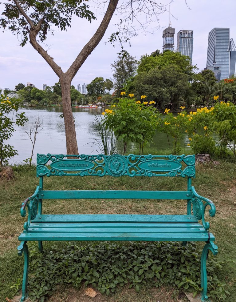 A decorative turquoise colour bench, situated in front of a lake, surrounded by trees and flowers, and city buildings in the background, in Lumphini Park, Bangkok, Thailand.