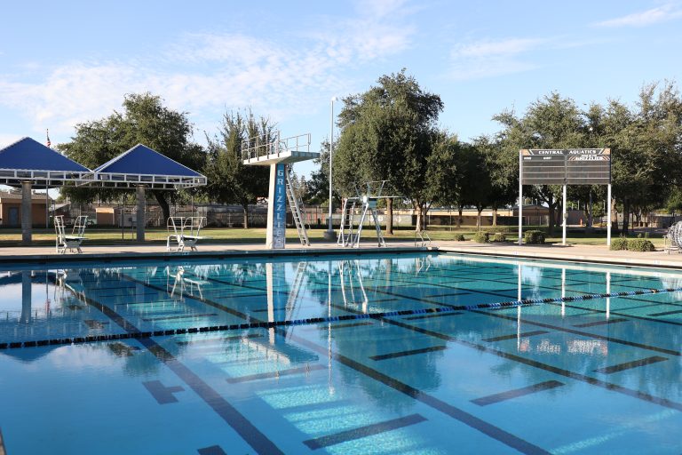 A clear outdoor swimming pool with lane dividers, surrounded by a concrete deck and a grassy area with trees.