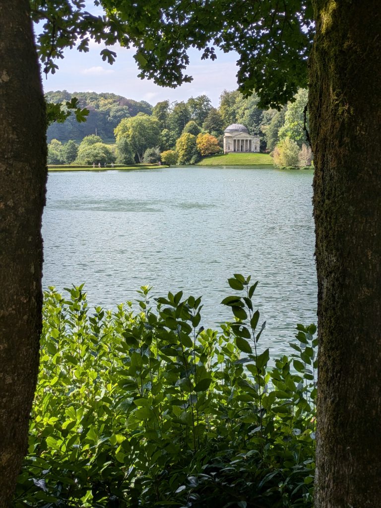 The view of the lake across to the Temple from the entrance garden side, at Stourhead National Trust gardens, in Wiltshire, England. In the photo the lake is framed by two tall and thick trees.