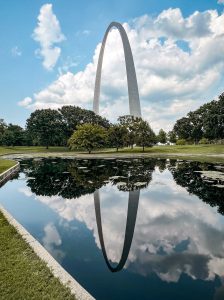 The image shows a reflective pond in a park with neatly trimmed grass and tall trees. The tall, stainless-steel st louis gateway arch rises in the background, and its reflection is clearly visible in the still water of the pond. The sky is bright with scattered clouds, adding to the serene atmosphere.