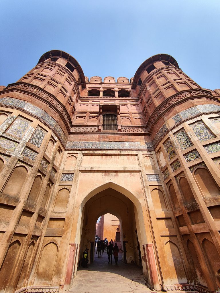 Entrance to agra fort, featuring towering bastions and intricate mughal architecture. The reddish-brown stone structure with decorative tilework and arched windows stands tall against a clear blue sky, with people passing through the grand arched gateway.