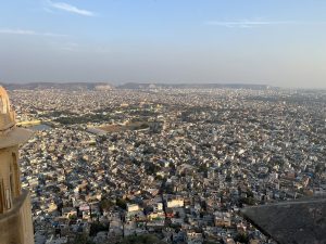 A panoramic view of Jaipur city from Nahargarh Fort, with a sprawling landscape of densely packed buildings stretching far into the horizon. The distant Aravalli hills form a natural backdrop under a clear blue sky. In the foreground, part of the fort's structure is visible, highlighting its elevated vantage point.