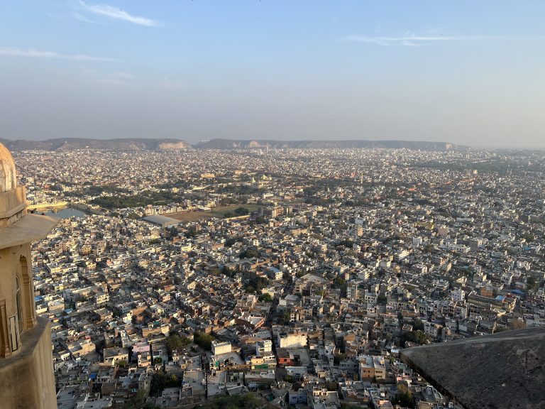 A panoramic view of Jaipur city from Nahargarh Fort, with a sprawling landscape of densely packed buildings stretching far into the horizon. The distant Aravalli hills form a natural backdrop under a clear blue sky. In the foreground, part of the fort’s structure is visible, highlighting its elevated vantage point.