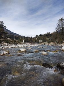 View larger photo: A clear river flows over smooth rocks, surrounded by trees and greenery, with snow-capped mountains in the background and a cloudy sky above. Gadherni, Himachal Pradesh.