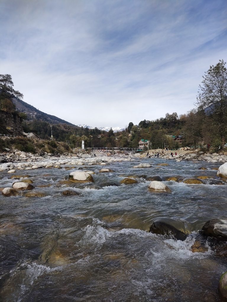 A clear river flows over smooth rocks, surrounded by trees and greenery, with snow-capped mountains in the background and a cloudy sky above. Gadherni, Himachal Pradesh.