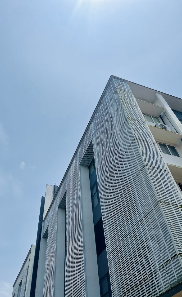 The corner of a modern building, photographed from below at a steep upwards angle. One side of the building is covered in perforated metal plating.